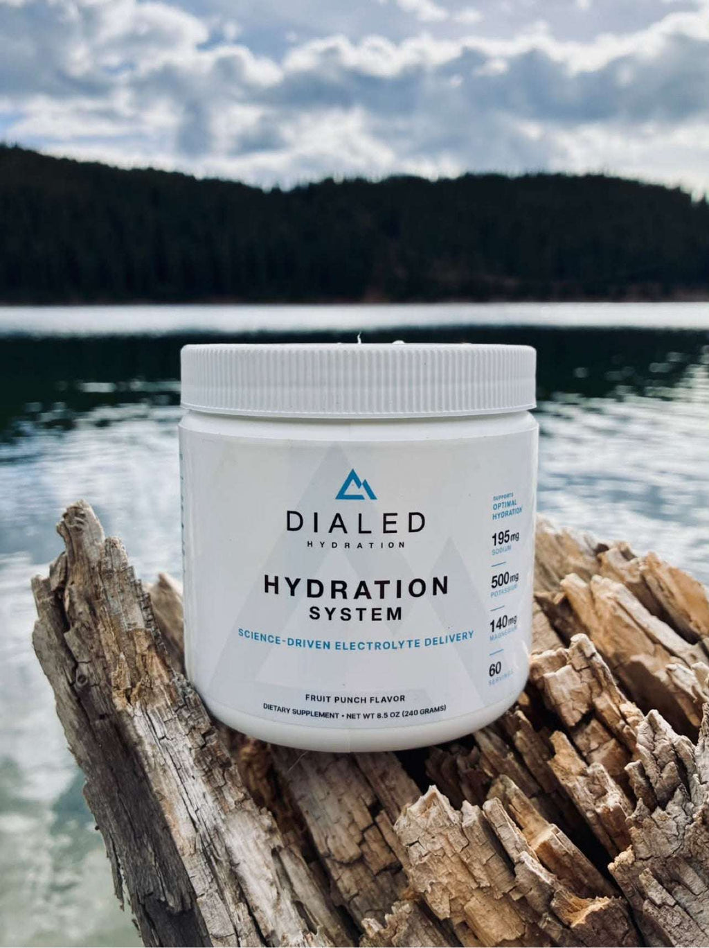 Dialed Hydration electrolyte drink mix in a white jar with fruit punch flavor, resting on driftwood beside a lake.