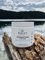 Dialed Hydration electrolyte drink mix in a white jar with fruit punch flavor, resting on driftwood beside a lake.